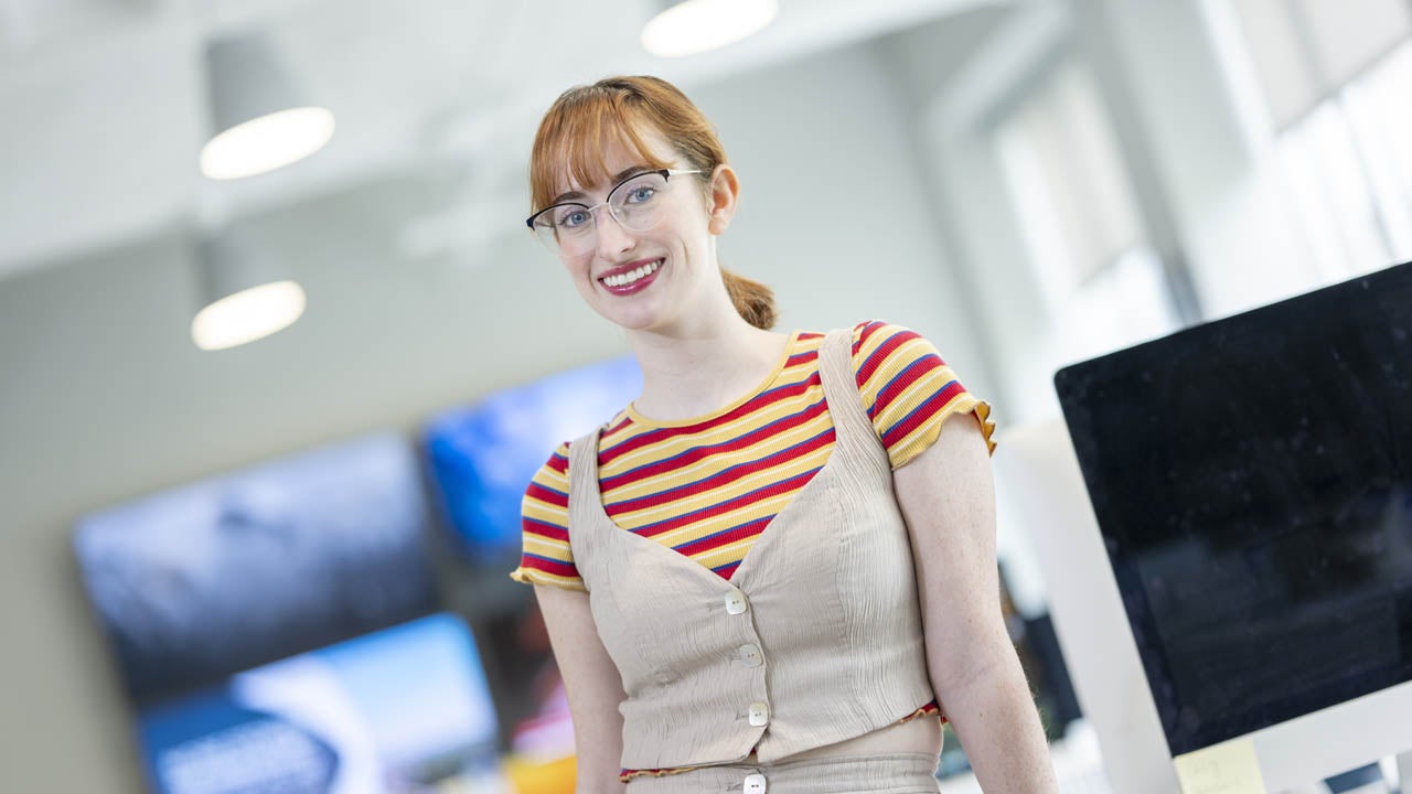 Student smiles while standing beside a desktop computer in a campus technology lab.