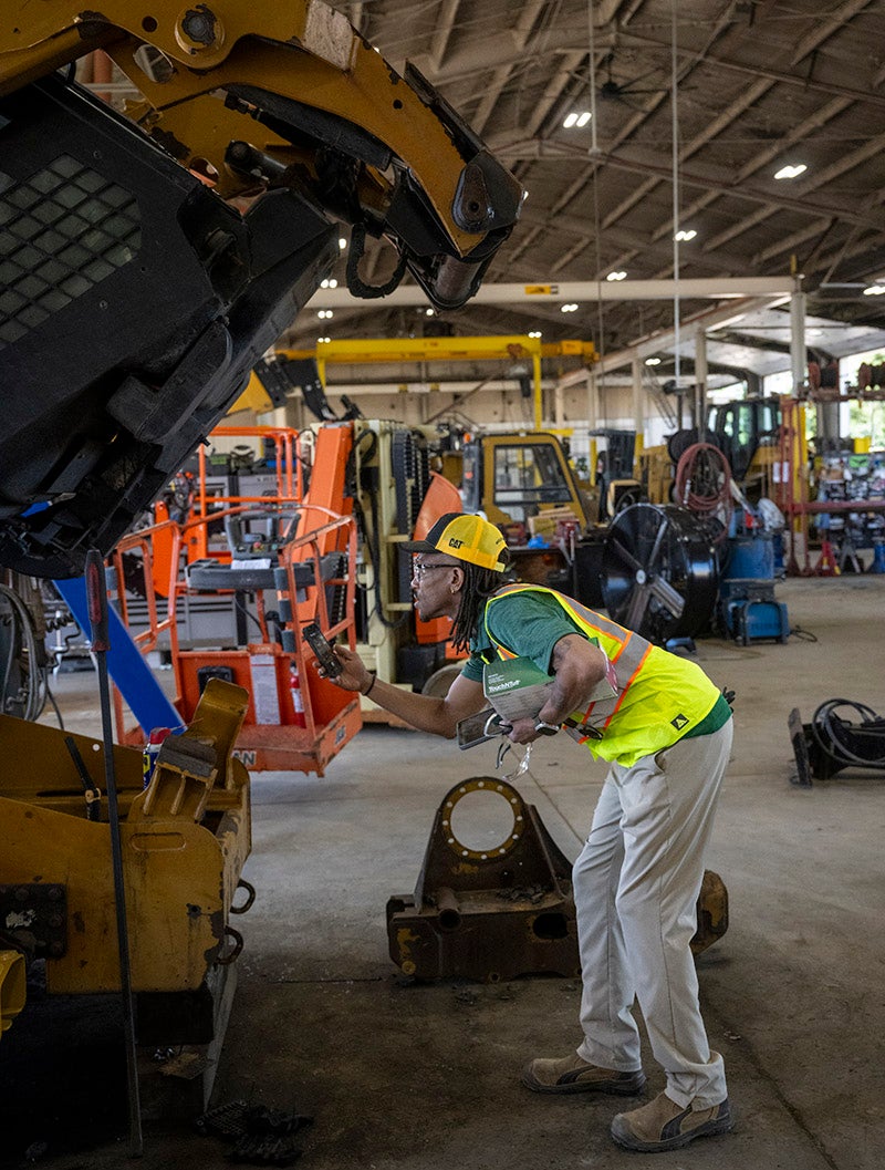 A person wearing a high-visibility safety vest, work boots, and a yellow CAT cap inspects a large piece of construction equipment inside a maintenance workshop. The person holds a handheld device while examining the raised machine. Additional heavy equipment, tools, lifts, and industrial work areas are visible throughout the facility.