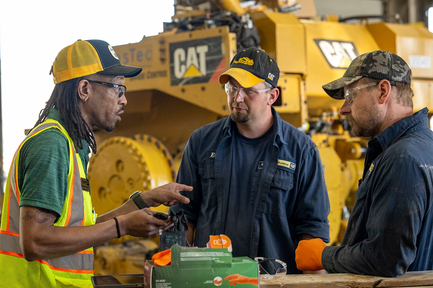 Three people wearing safety glasses stand beside large CAT equipment in a maintenance workshop. One person in a high-visibility safety vest holds a pair of work gloves while speaking with two people in work uniforms. Boxes of protective gloves and safety gear are visible on a work surface in the foreground.