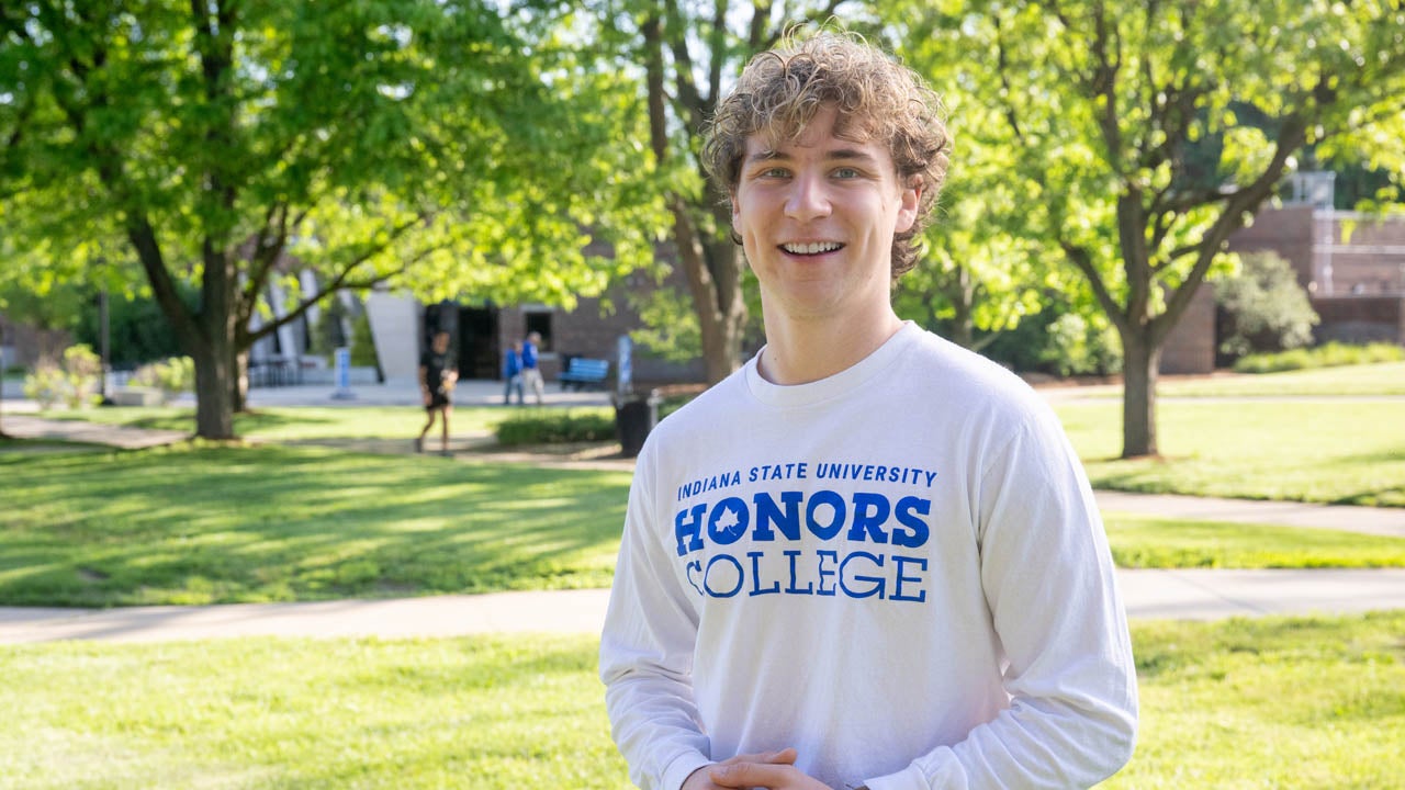 Student wearing an Indiana State University Honors College shirt stands outdoors on a tree-lined campus.