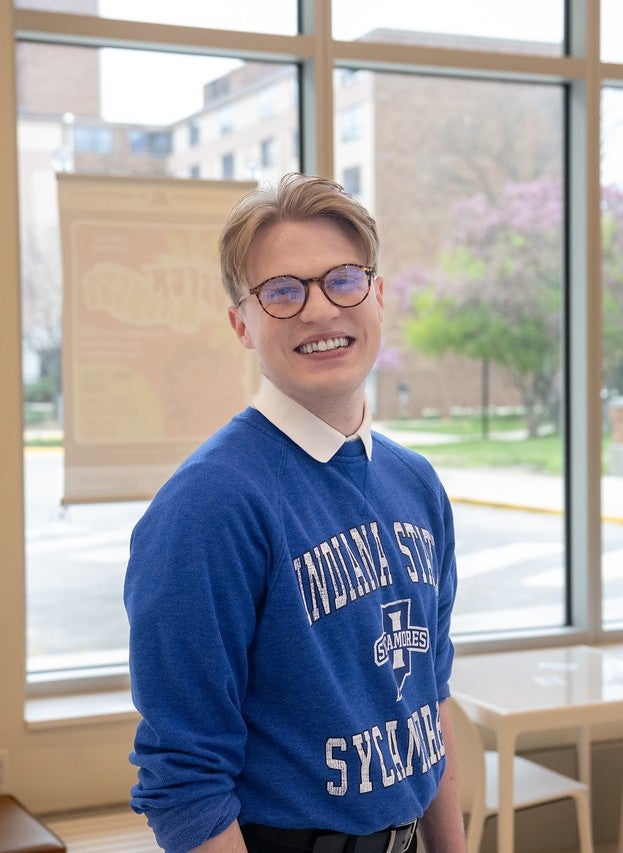 Person wearing a blue Indiana State Sycamores sweatshirt and glasses stands in a campus building with large windows, tables, and chairs in the background.