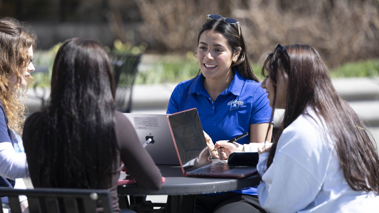 Students work together on laptops while seated around an outdoor table on campus.