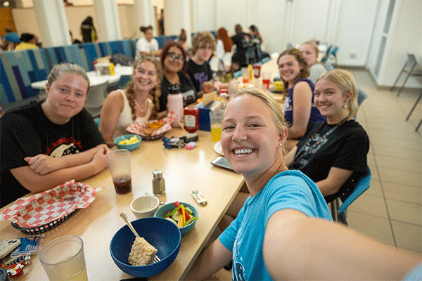 Group of students seated around a long table in a campus dining area. One smiling student holds a camera toward the group for a selfie while trays, bowls, drinks, and condiments are spread across the table.