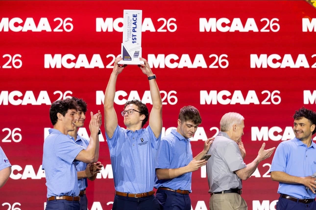 Several people wearing matching light blue polo shirts stand on a stage in front of a red backdrop displaying “MCAA 26.” One person at the center holds a first-place trophy overhead while others nearby applaud.