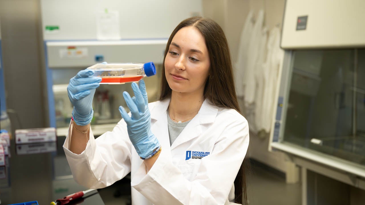Student wearing a lab coat and gloves examines a cell culture flask in a laboratory.