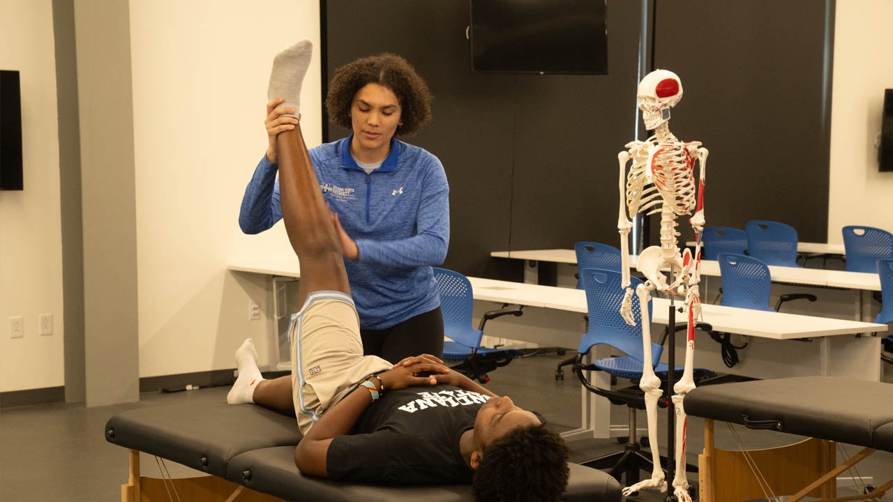 A student lies on a treatment table while an instructor stretches their leg in a classroom with an anatomical skeleton model.