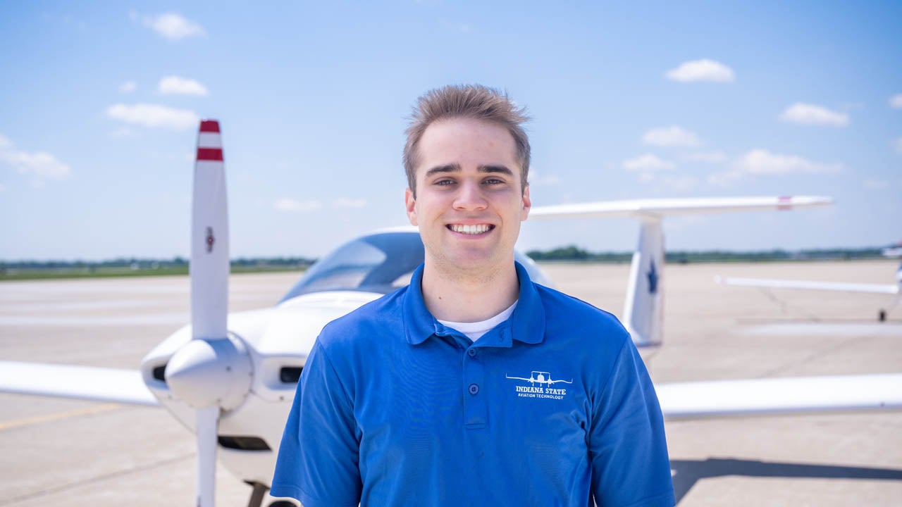 Student wearing an Indiana State University Aviation Technology polo stands in front of a small airplane on an airport tarmac.