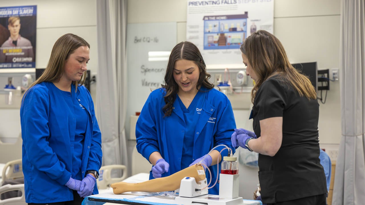 Nursing students practice a clinical procedure on a training mannequin arm in a simulation lab.