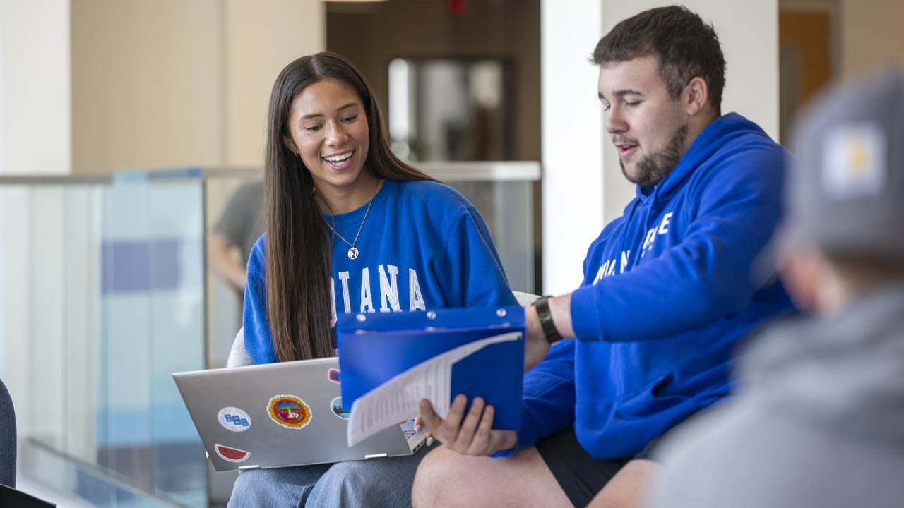 Two students review materials together on a laptop and in a binder while seated in a campus study area.