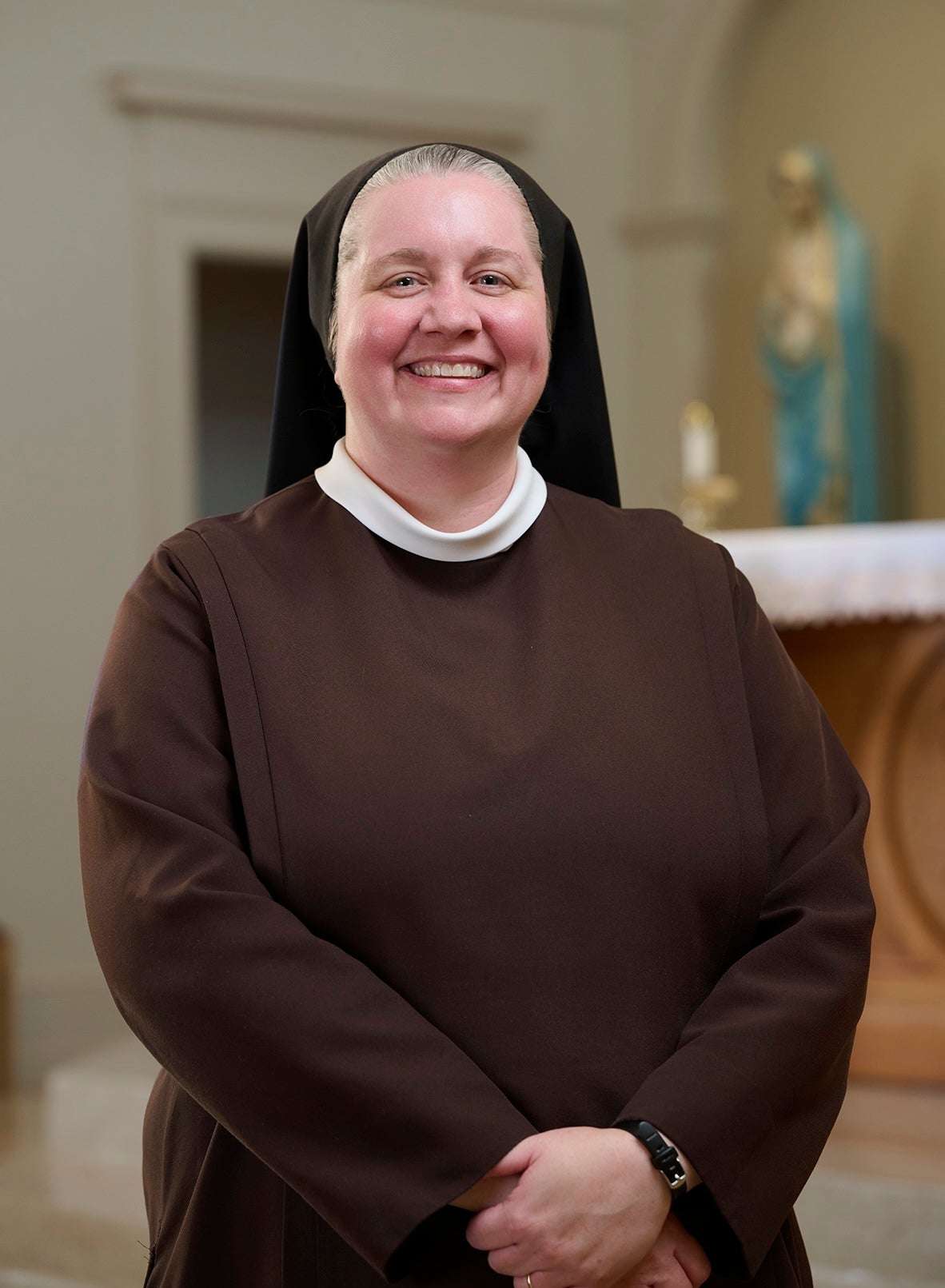 Sister Maria Gemma Salyer, OSF, smiles for a portrait while wearing a brown religious habit and black veil. She stands with her hands clasped in front of her inside a chapel.