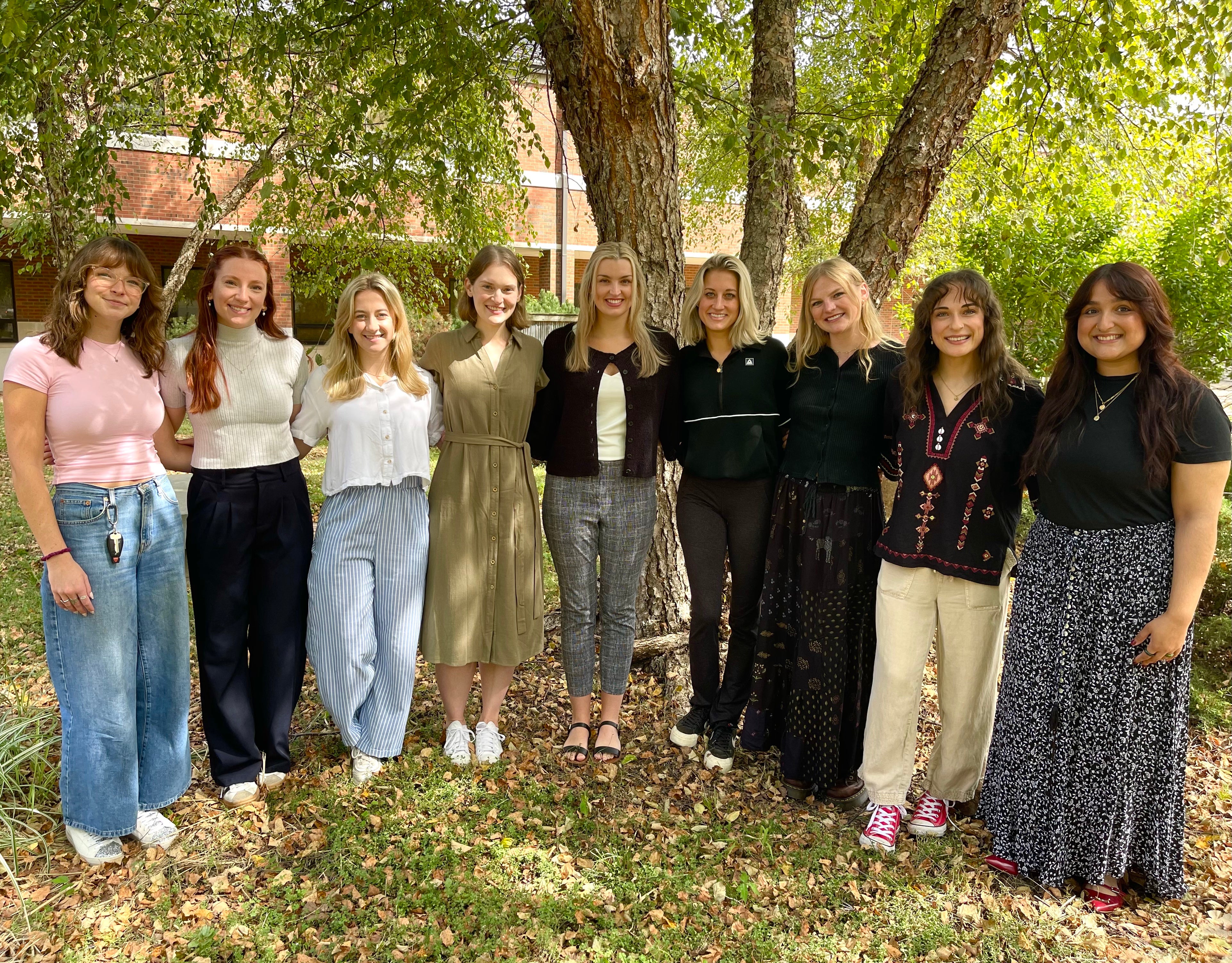 Nine people stand side by side outdoors beneath large trees, posing for a group photo on a grassy area with a brick campus building in the background.