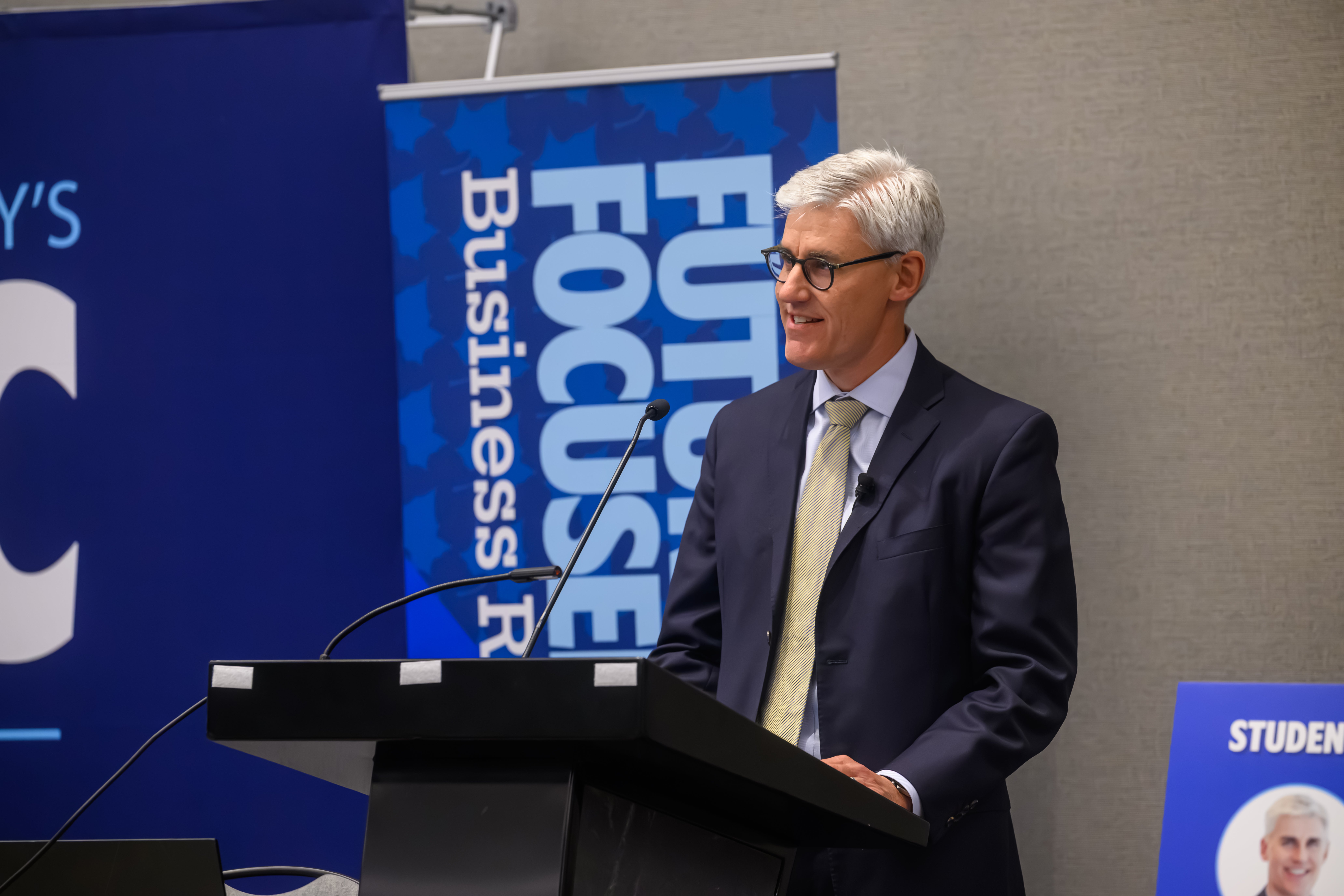 A person in a suit stands at a podium speaking, with a banner behind displaying "Future Focused Business".