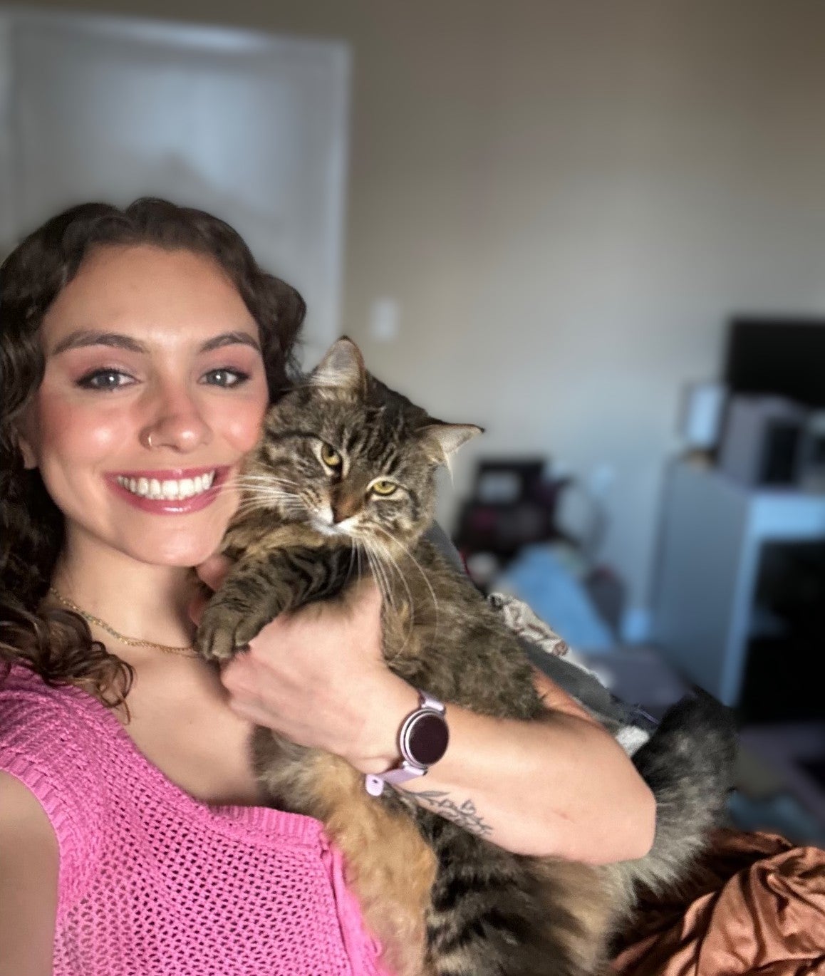 Selfie of a smiling woman with wavy brown hair, warm makeup, and a pink patterned top, holding a fluffy brown tabby cat close to her cheek.
