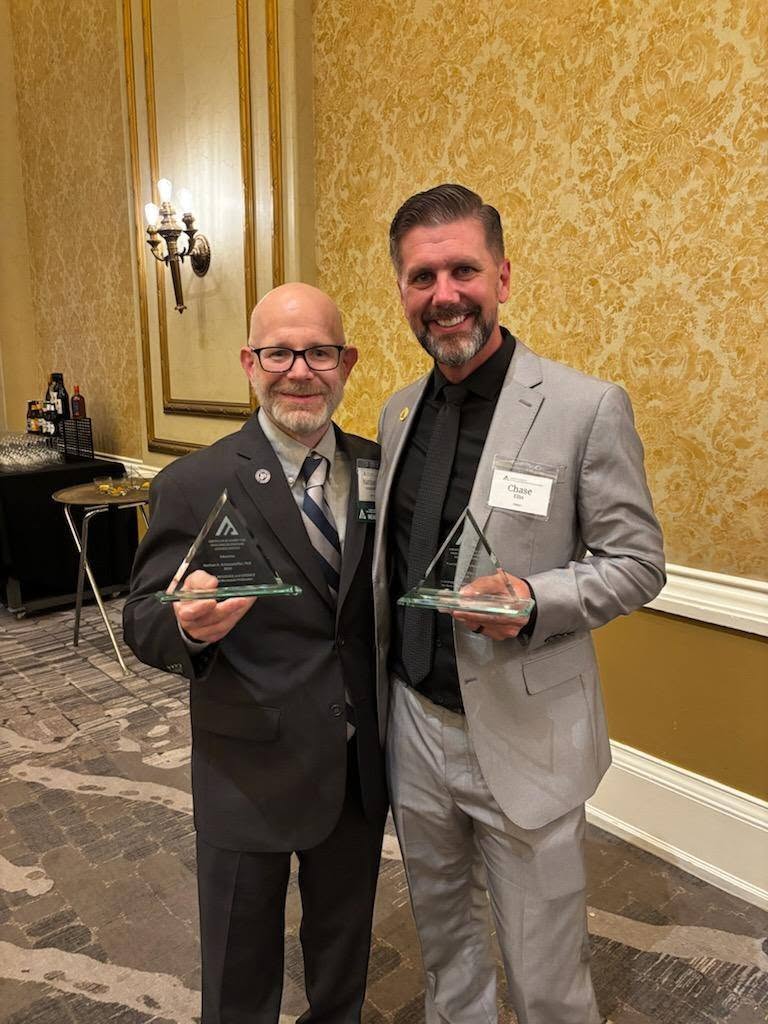 Two individuals (Dr. Schaumleffel, left and Chase Ellis, right) are standing in a room with ornate wallpaper, holding clear triangular awards. They are both dressed in formal suits with name tags. 