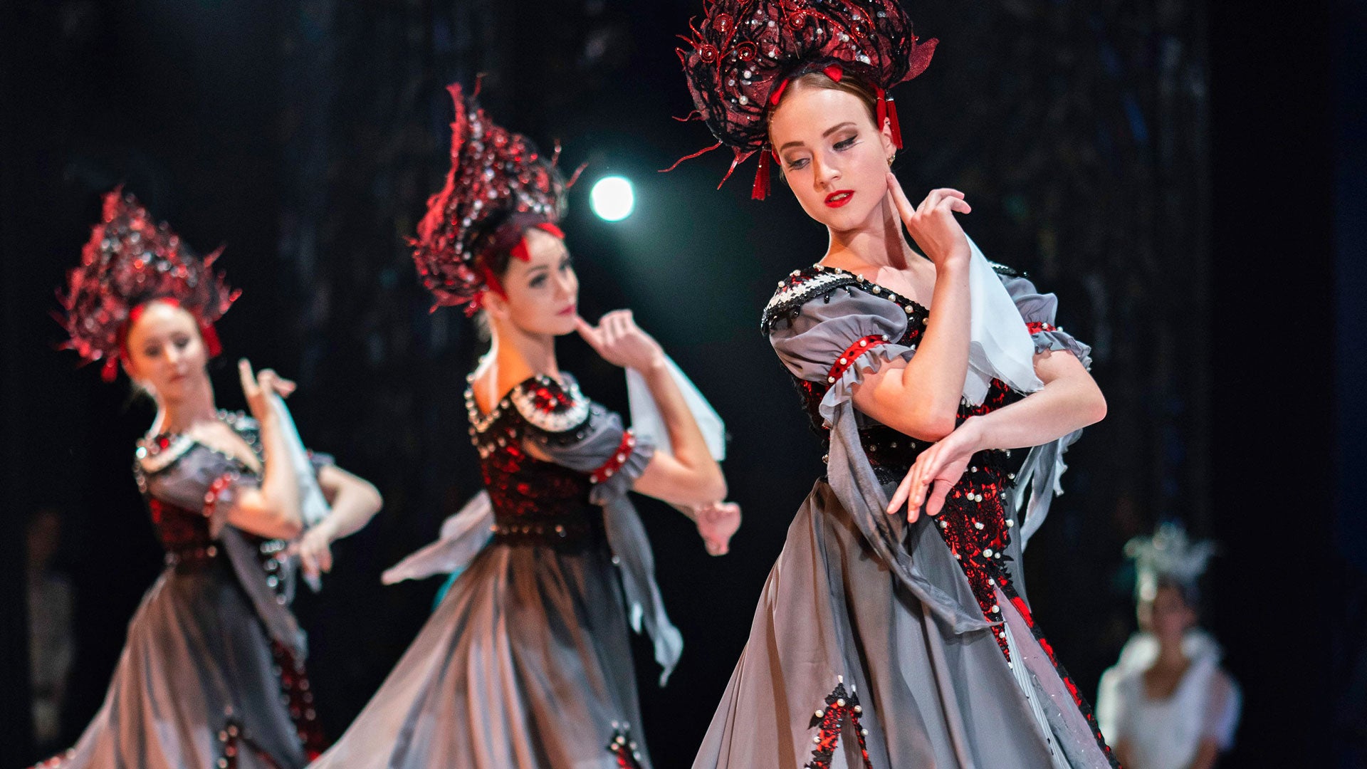 Three ballet dancers on stage in grey and red dresses with elaborate, dark ornamental headdresses, posing with graceful hand gestures.