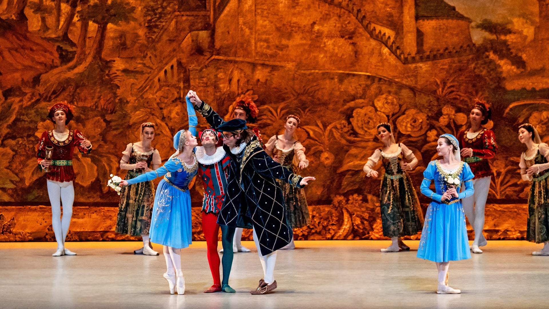A group of ballet performers on stage in Renaissance-style costumes, featuring a central jester in a red and blue costume flanked by dancers in blue dresses and a nobleman in a dark coat, against a painted golden-brown backdrop.