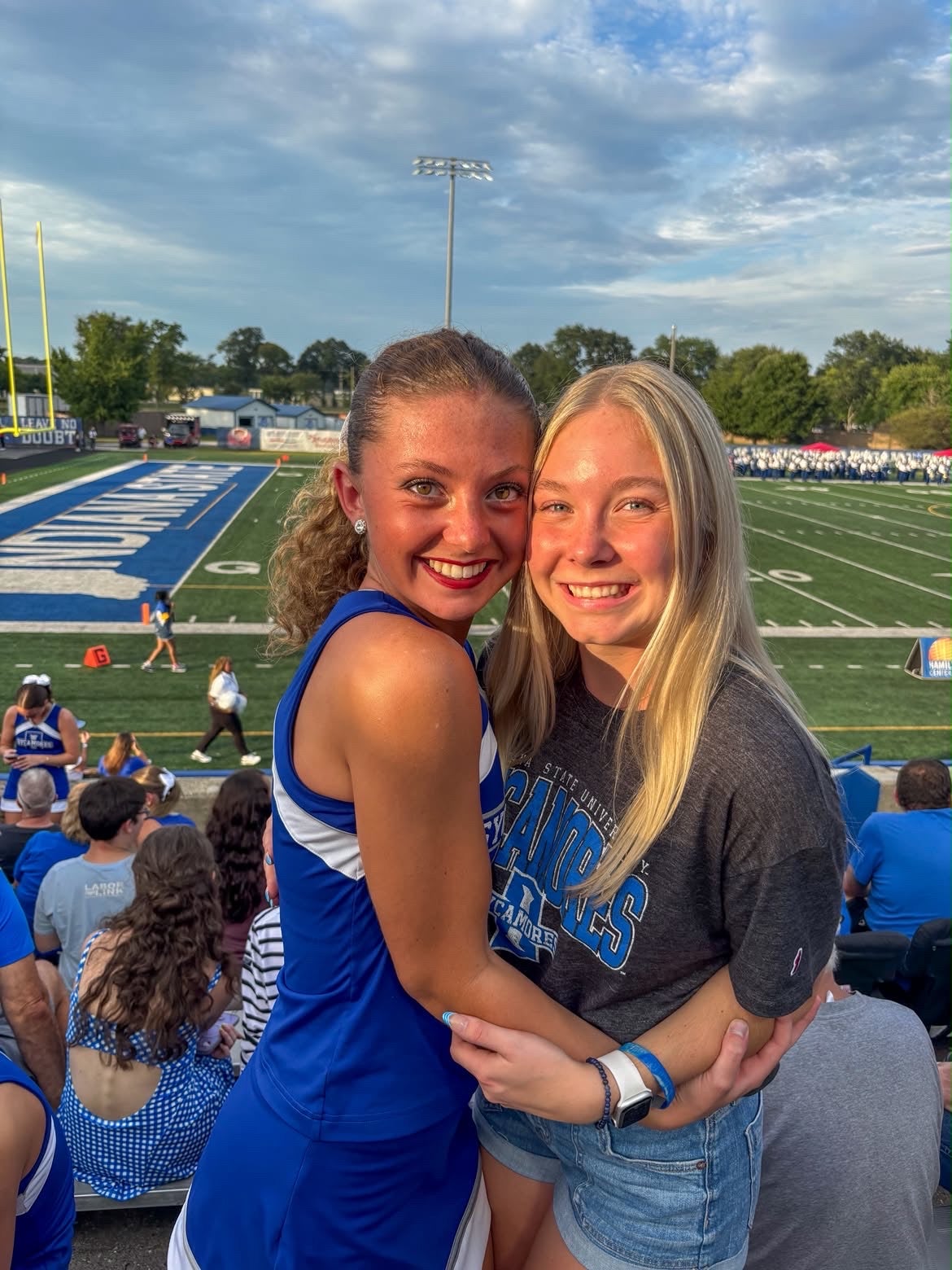 Two young women stand together in the bleachers at a football stadium, posing in front of the field during an outdoor event. One wears a blue athletic uniform, and the other wears a dark Indiana State University T-shirt and denim shorts. Spectators, the football field, stadium lights, and a marching band are visible in the background.