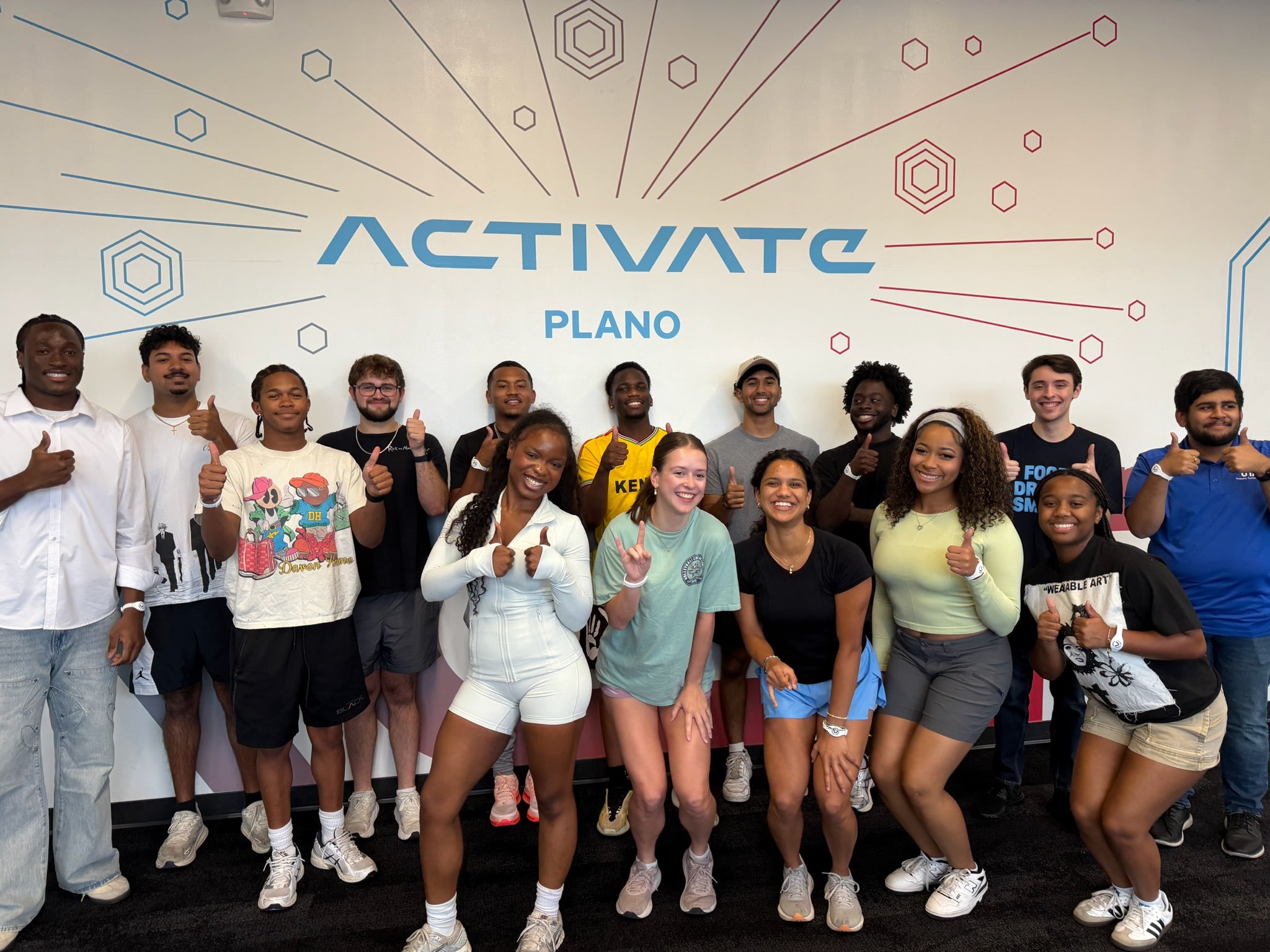 A group of students poses together, many giving a thumbs-up, in front of a wall displaying the words “Activate Plano.”
