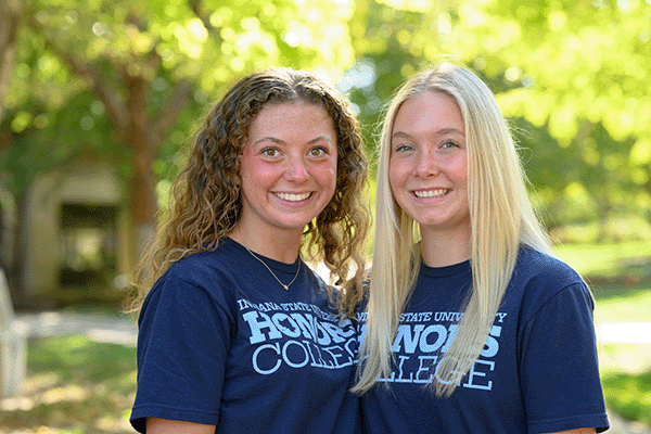 Two people standing side by side outdoors, wearing matching navy blue Indiana State University Honors College T-shirts. Trees and a campus building are softly blurred in the background.