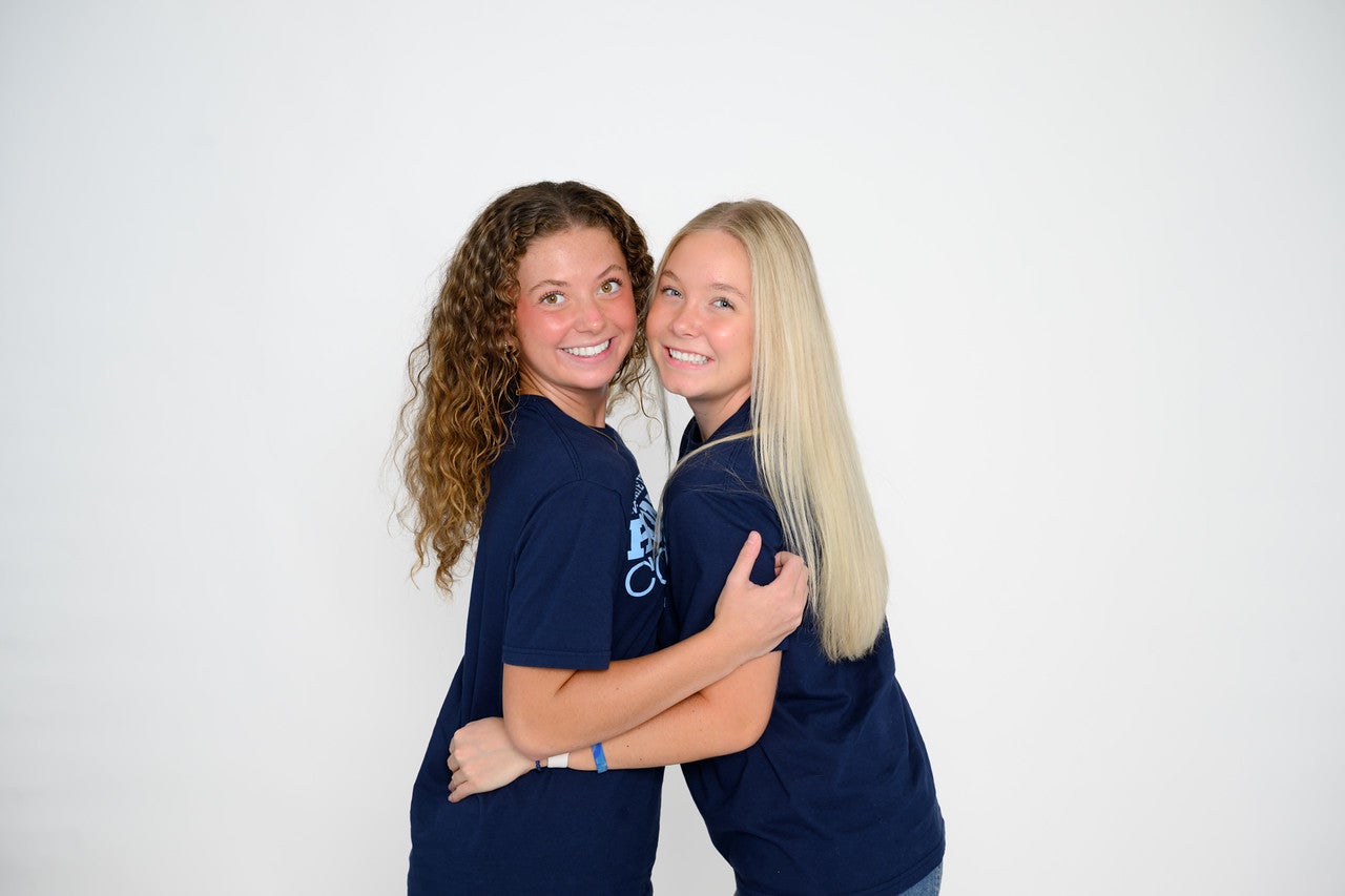 Two young women wearing matching Indiana State University Honors College T-shirts stand together against a plain light-colored studio background. The pair face the camera with their arms around each other in a posed portrait.