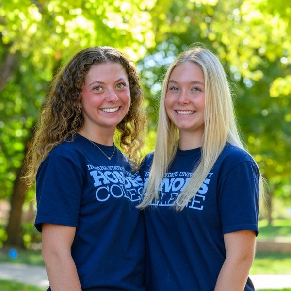 Two young women wearing matching Indiana State University Honors College T-shirts stand side by side outdoors beneath leafy trees. The photo is framed from the waist up, with a sunlit green background and soft-focus foliage surrounding the subjects.