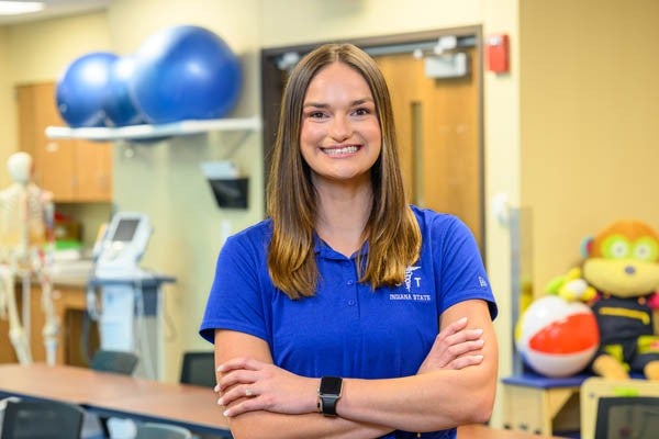 Female with brunette hair wearing an Indiana State blue polo stands in a classroom with arms crossed, with therapy equipment, exercise balls, a skeleton model, and adaptive toys in the background.