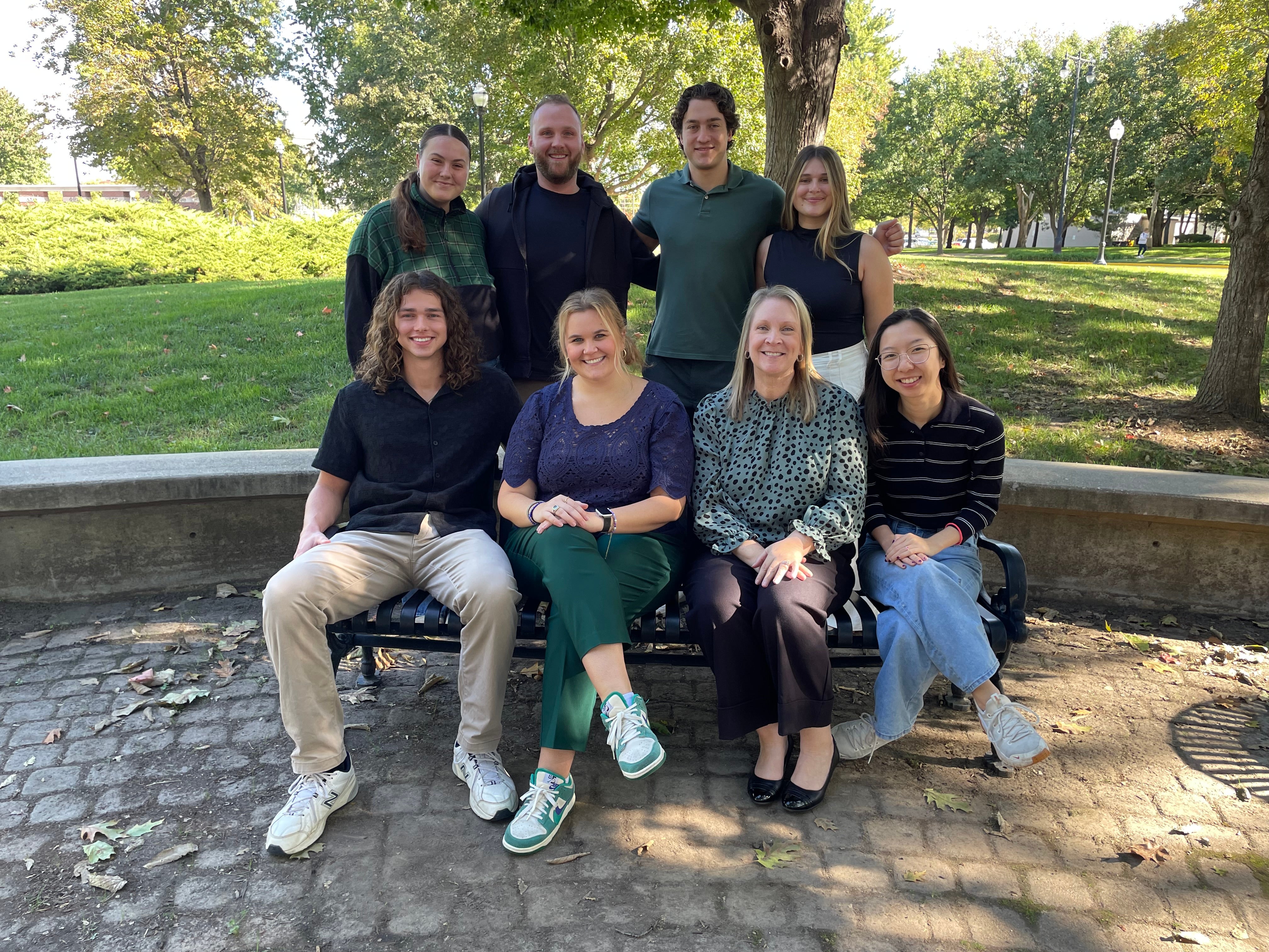Group photo of eight students and faculty members posing outdoors on a sunny day. Four individuals are seated on a park bench over cobblestone, and four are standing behind them, with trees, a grassy lawn, and park lamps visible in the background.