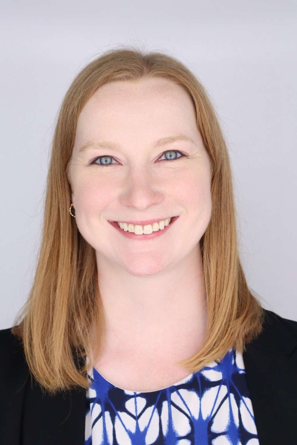 Professional headshot of a woman with shoulder-length blonde hair, pale skin, and blue eyes, smiling warmly. She is wearing a black blazer over a blue and white patterned top.