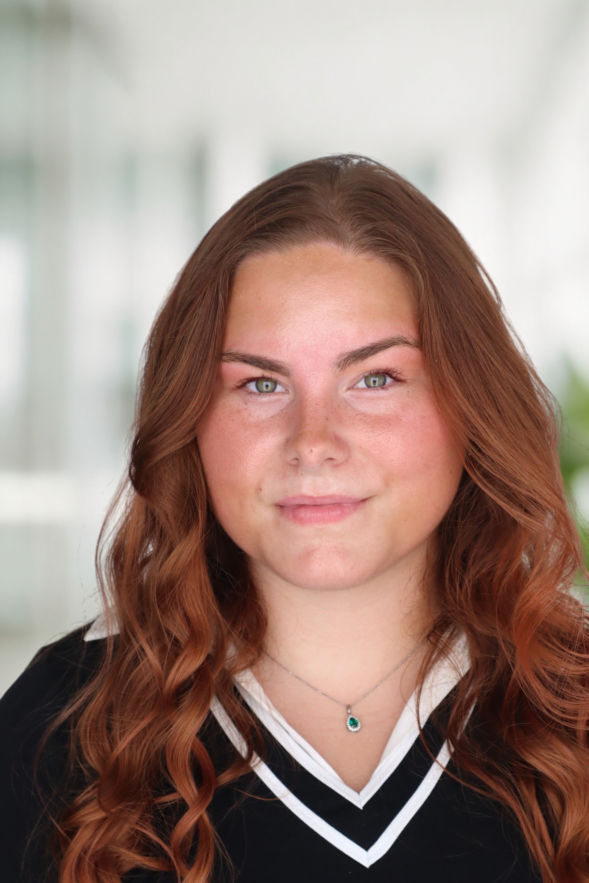 Professional headshot of a smiling young woman with long, wavy red hair, pale skin, freckles, and green eyes. She is wearing a black top with a white V-neck collar and a silver necklace with a green gemstone pendant.