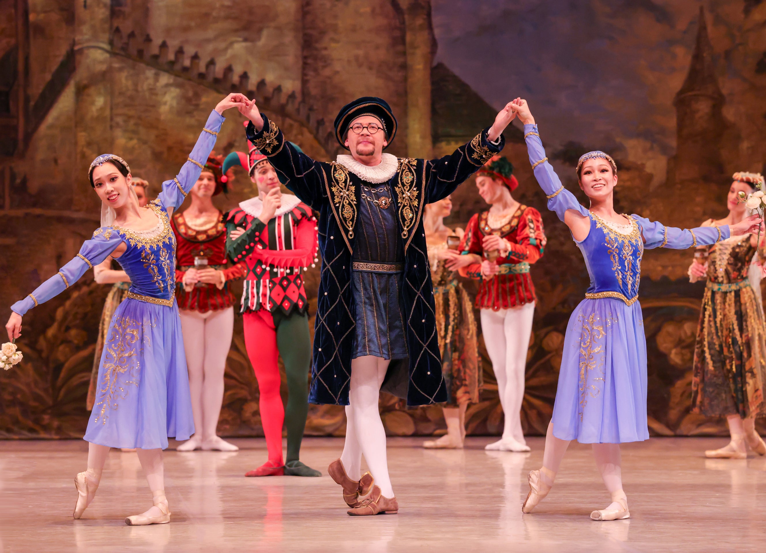 A central male performer in a dark blue velvet coat and beret holds hands with two female ballet dancers in matching blue and gold dresses, with an ensemble cast in historical costumes standing in the background against a painted castle backdrop.