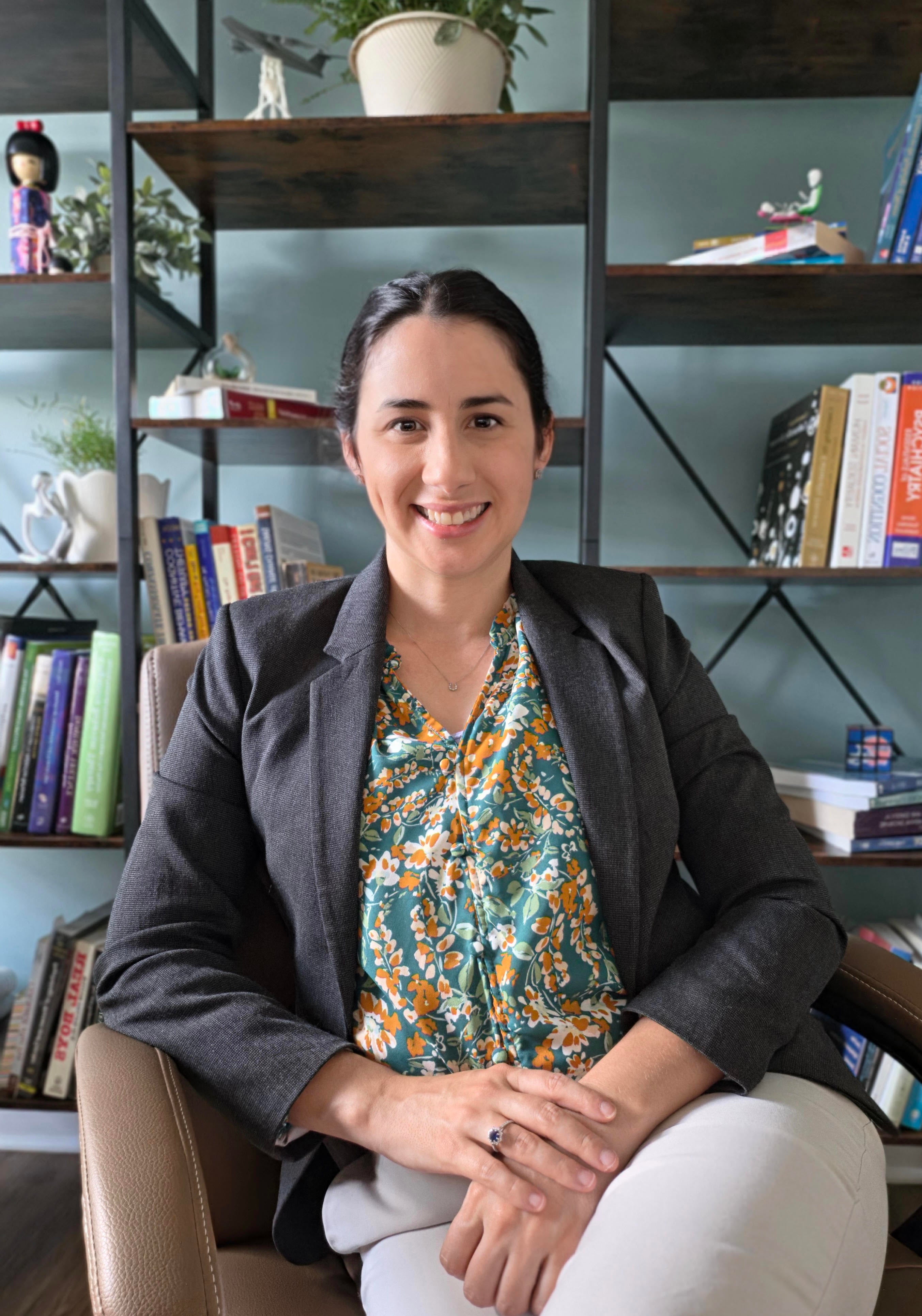 A professional headshot of a woman sitting in a chair in front of a bookshelf, wearing a dark gray blazer over a green and orange floral blouse, with her hands clasped in her lap and smiling at the camera.