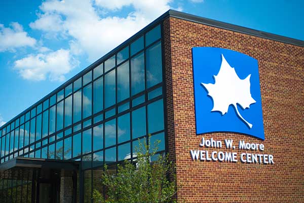 The John W. Moore Welcome Center is seen in the daytime, with blue sky and white clouds reflected in the building’s windows to the left. A large white Sycamore leaf on blue background is seen on the building’s red brick wall to the right, with the name underneath.