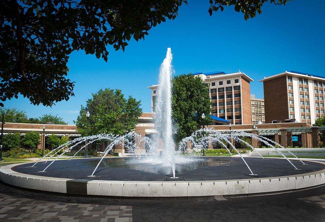 Large circular water fountain with multiple streams in front of Indiana State University buildings under clear blue sky.