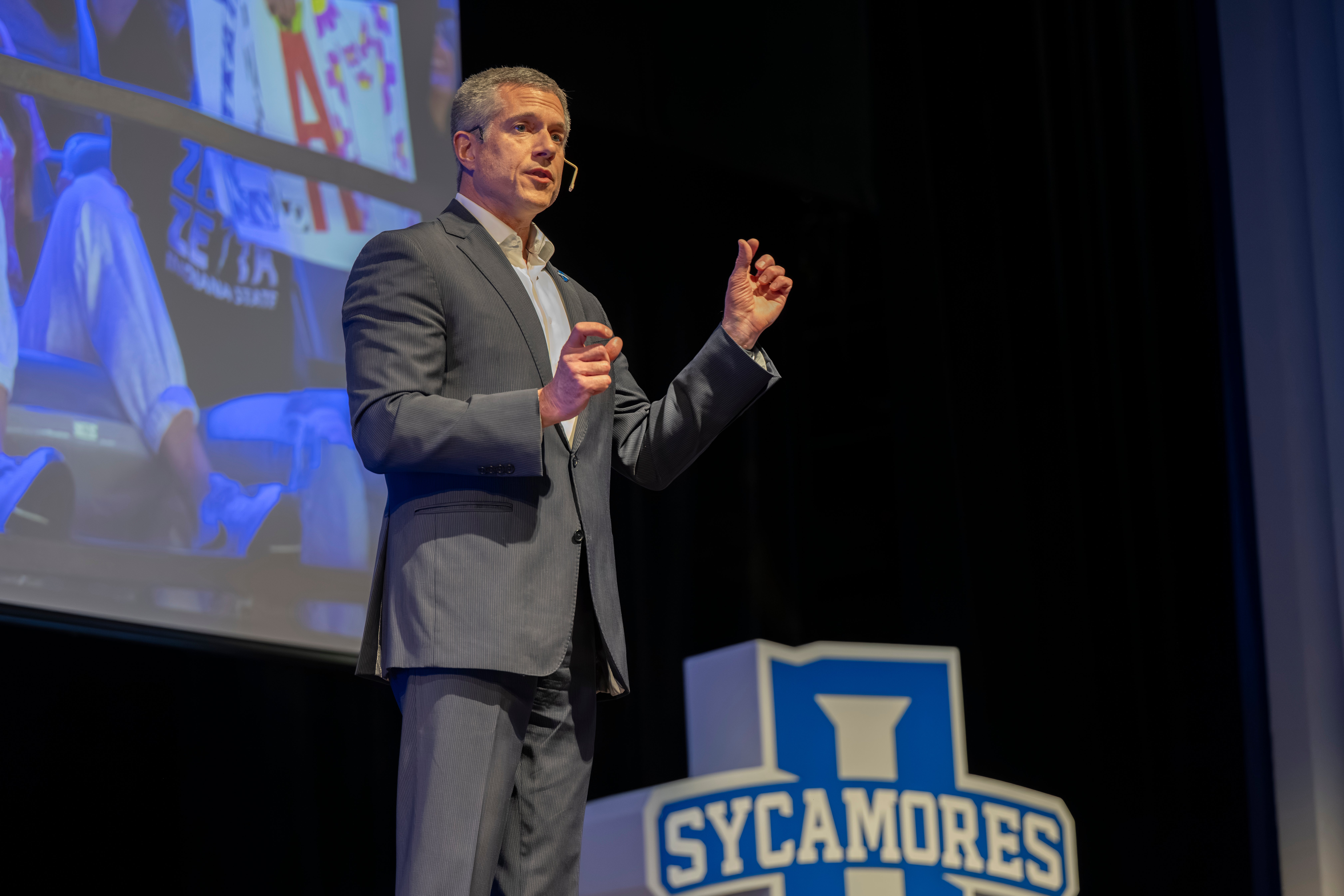 Man in gray suit speaking on stage with Sycamores logo visible near podium and a large screen behind him.