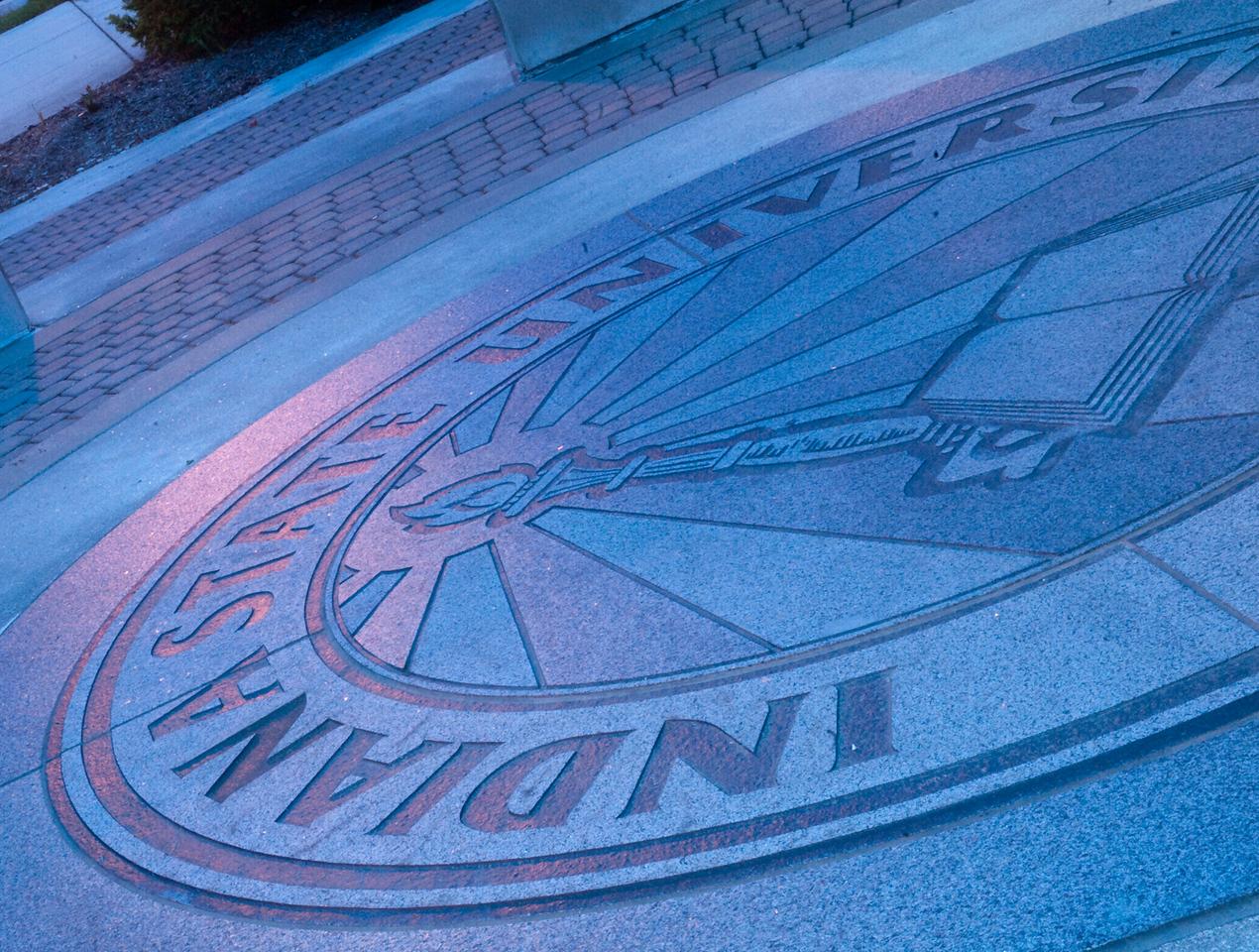Indiana State University circular emblem etched in stone on an outdoor walkway, featuring a torch and an open book.