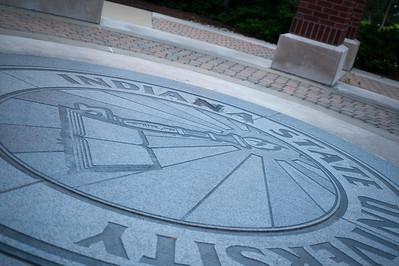 Engraved Indiana State University seal set in a circular stone pavement outdoors near a brick pillar.
