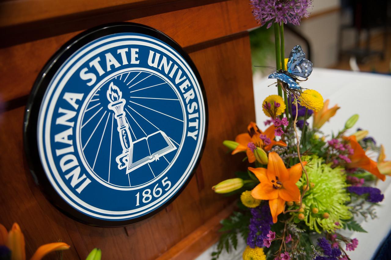 Indiana State University emblem on a wooden podium with a colorful floral arrangement featuring orange lilies and a decorative blue butterfly.