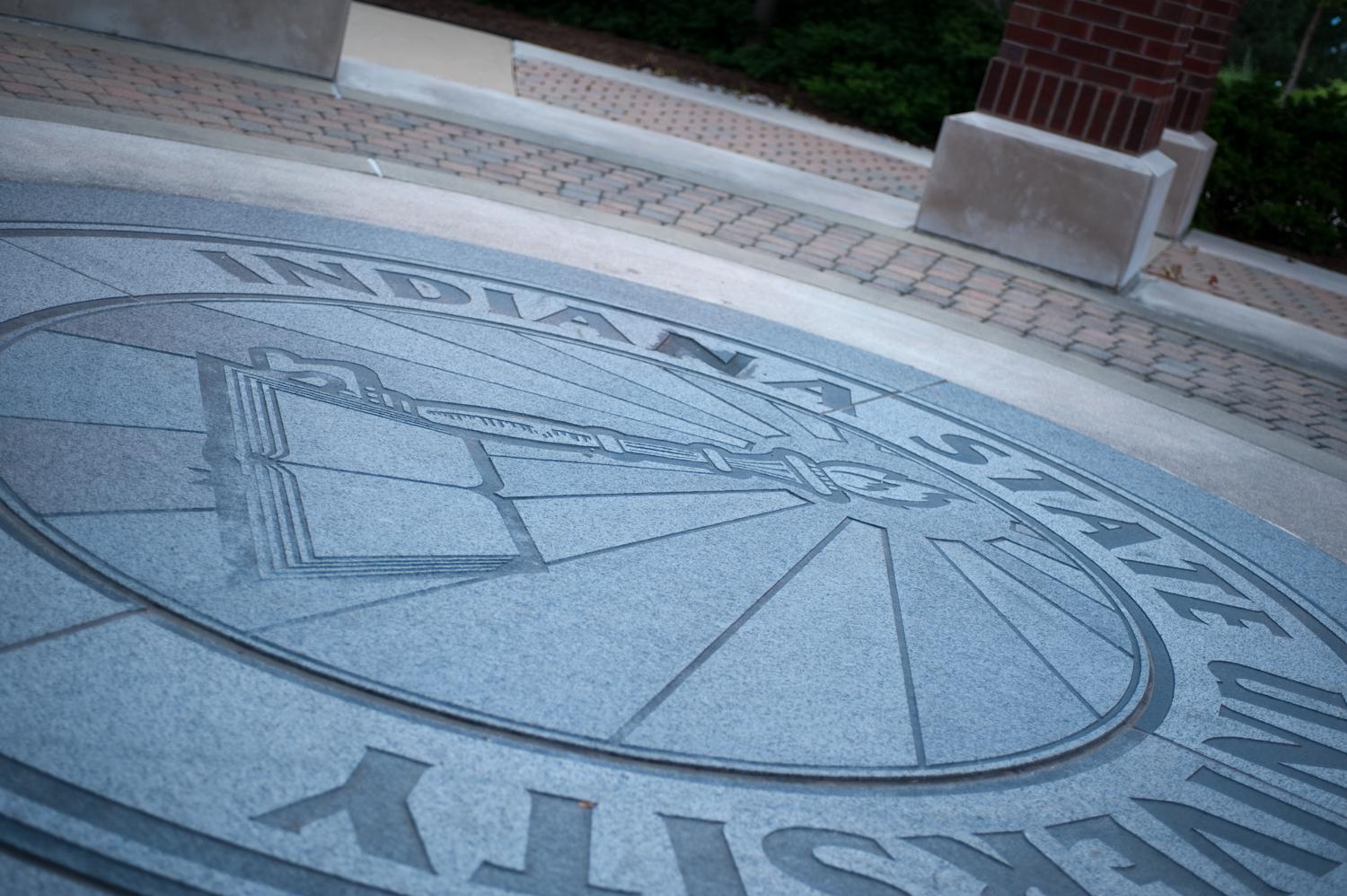 Indiana State University seal engraved in stone on the ground near brick columns and a paved walkway.