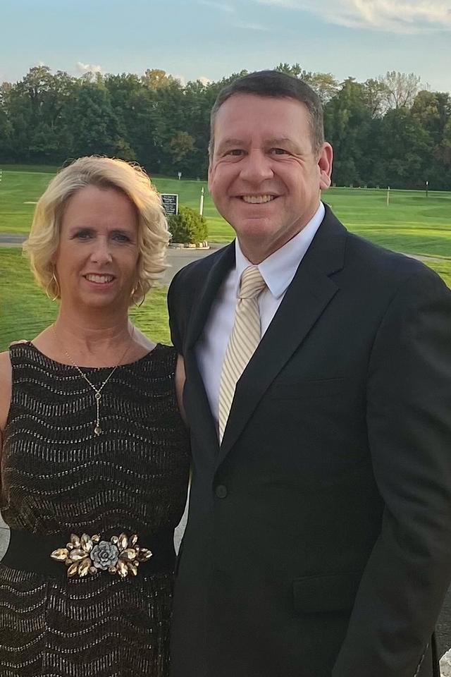 A man in a dark suit and light tie stands next to a woman in a black dress with a decorative belt, outdoors with trees and grass in the background.