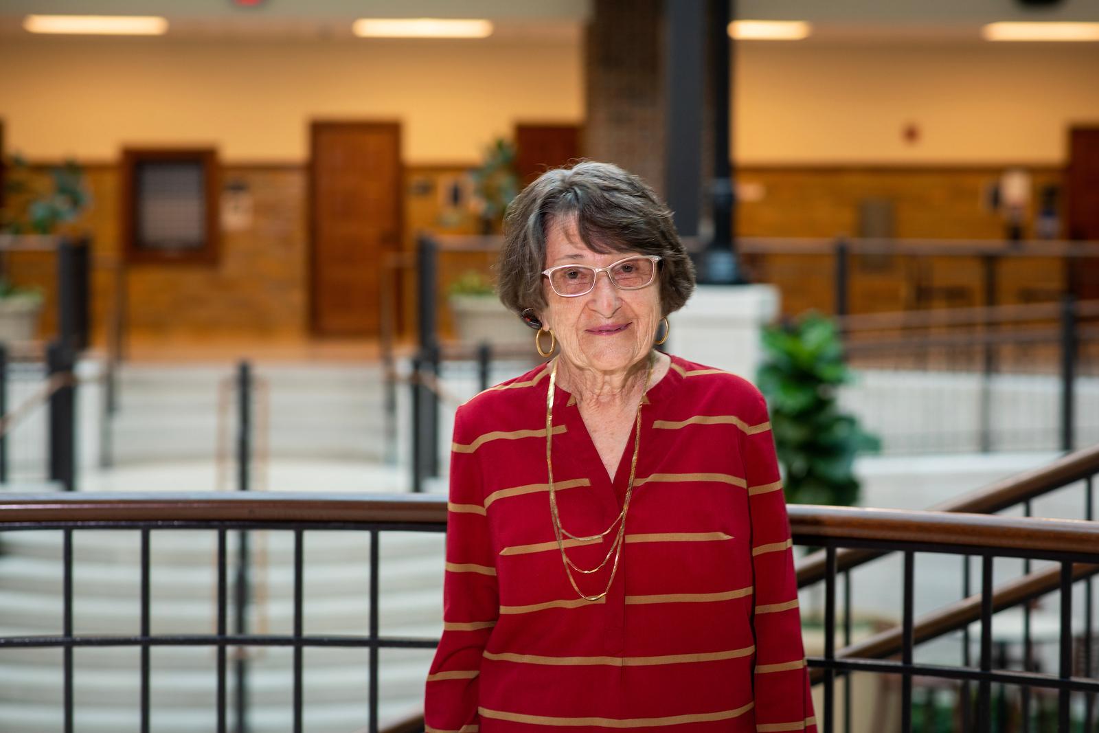 Elderly woman with short gray hair and glasses wearing a red blouse with yellow stripes, standing indoors near a metal railing.