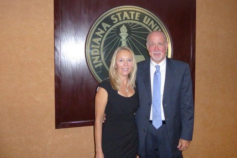 A man in a suit and woman in a black dress stand side by side, smiling, in front of an Indiana State University emblem on a wood-paneled wall.