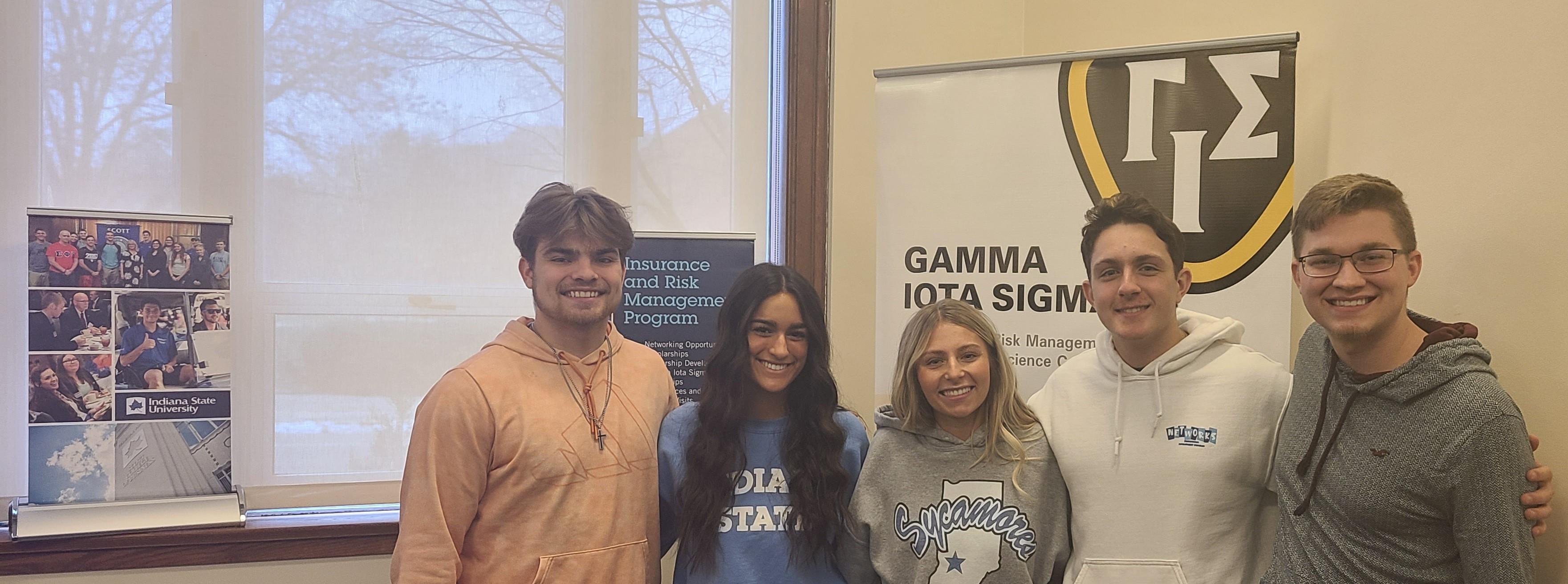 Five smiling college students stand indoors in front of two banners, one reading Gamma Iota Sigma and the other Indiana State University Insurance and Risk Mana