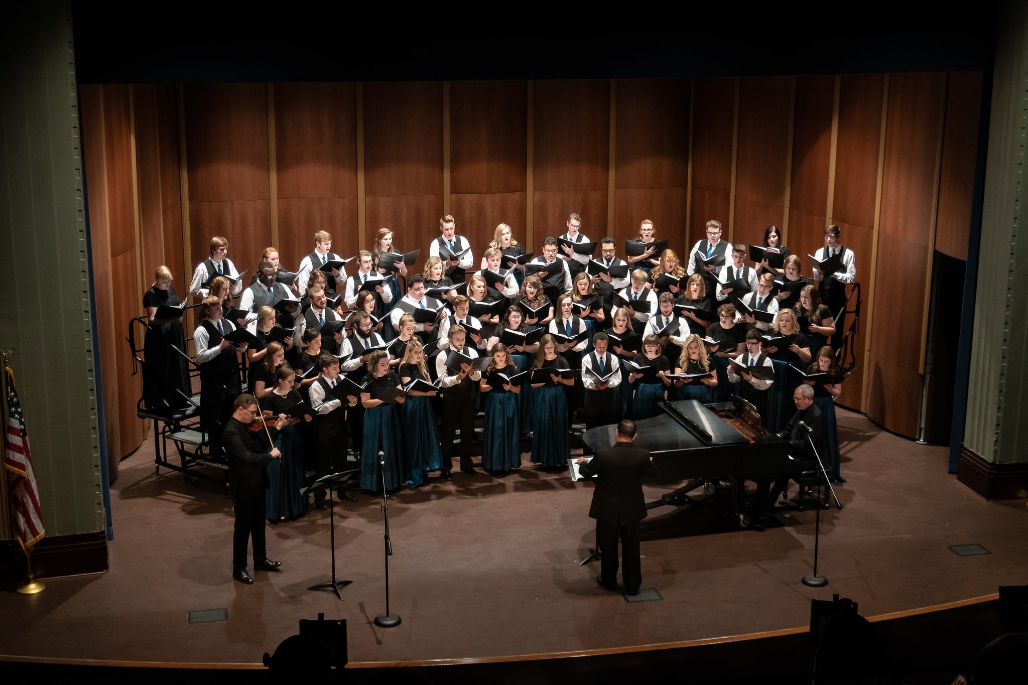 Large mixed choir performing on stage with a conductor, a violinist, and a pianist, all dressed formally, with wooden paneling backdrop.