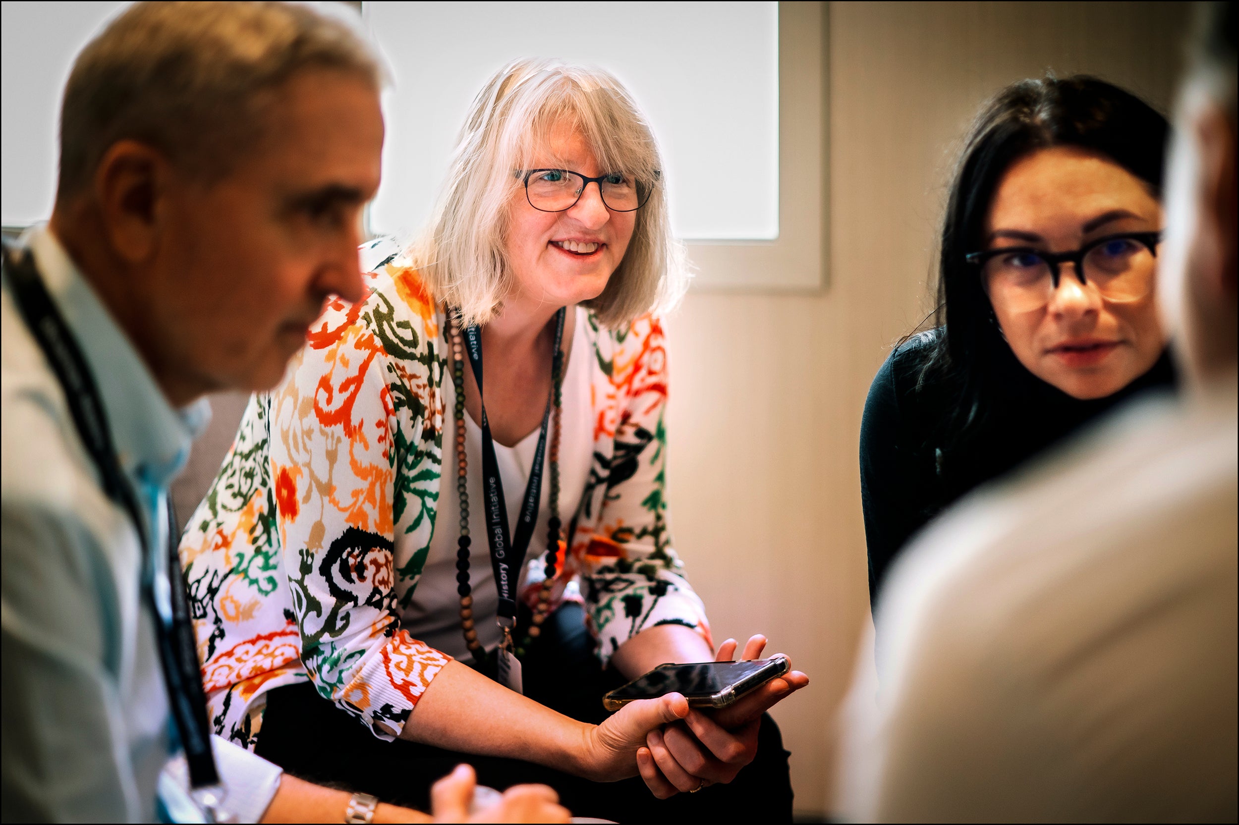 Four people engaged in discussion, one woman with glasses holding a phone and wearing a colorful patterned shirt smiling, others listening attentively.