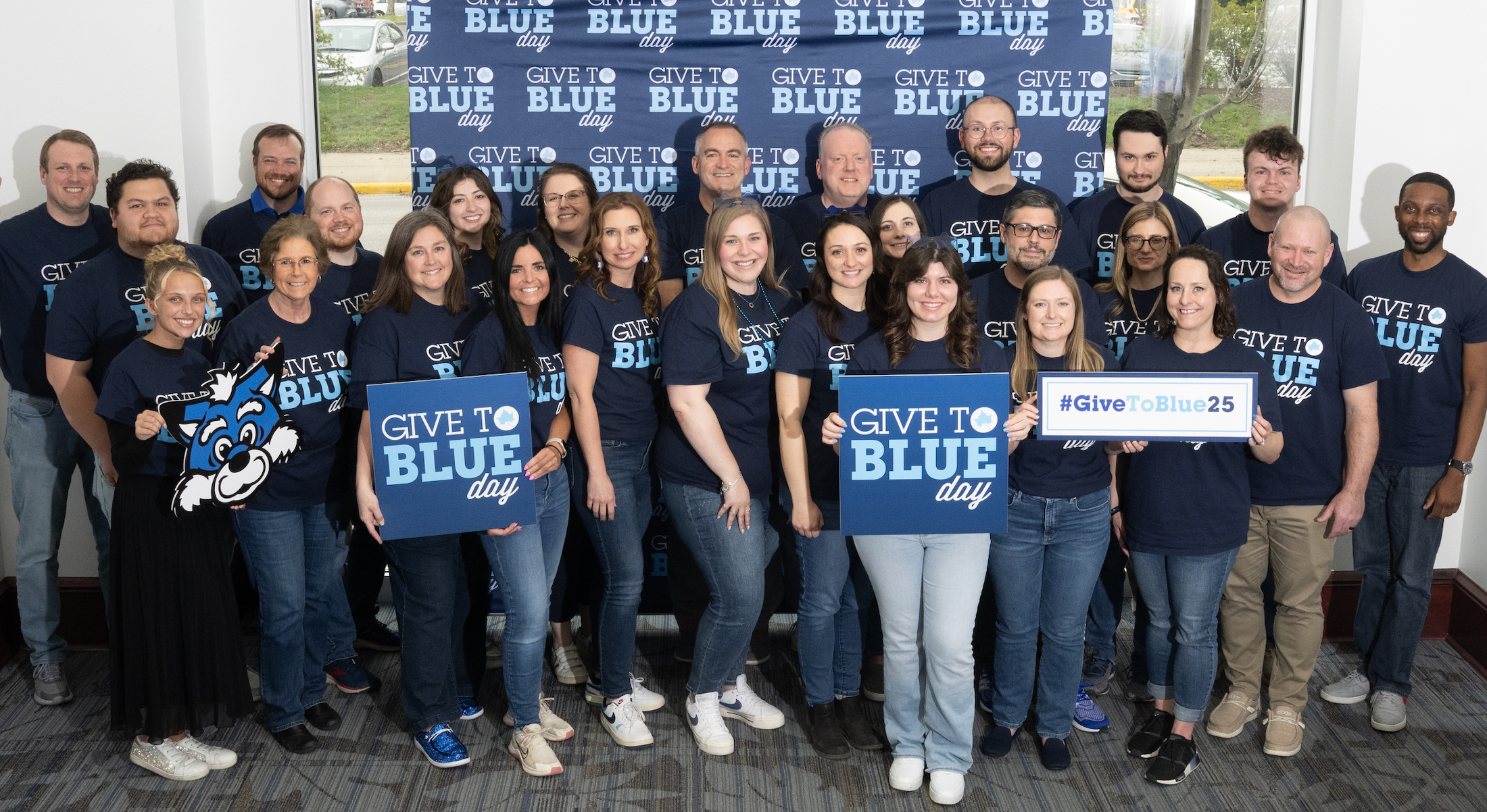 Group of diverse people wearing navy blue Give to Blue Day t-shirts, holding signs and mascot cutout, smiling in front of a Give to Blue Day backdrop.