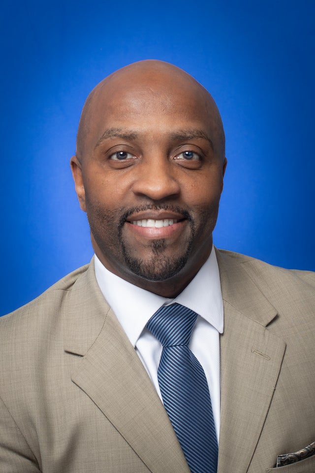Smiling man with a shaved head and goatee wearing a tan suit jacket, white shirt, and blue striped tie against a blue background.