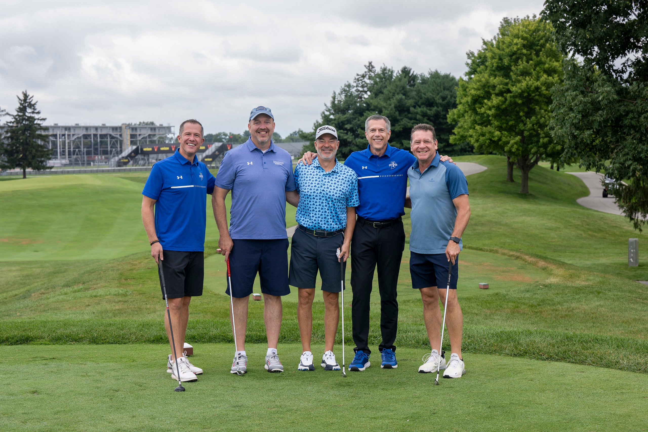 Five men standing on a golf course fairway, smiling, dressed in golf attire, each holding a golf club, with trees and a stadium in the background.