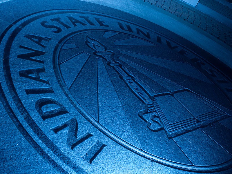 Indiana State University seal embedded in pavement, featuring a torch and open book, illuminated with blue lighting.