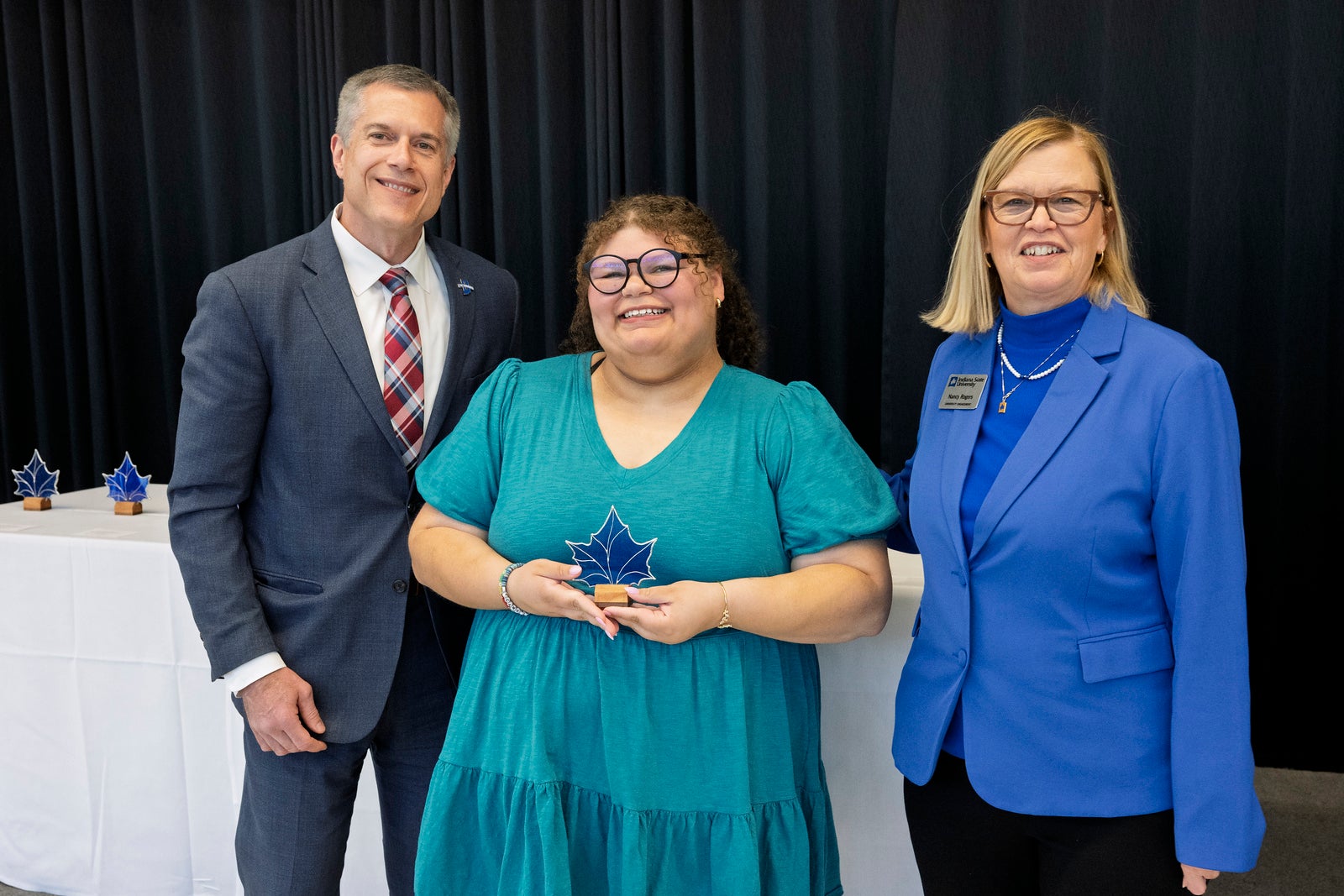 Three people smiling at awards ceremony; woman in center holding blue maple leaf-shaped award; man in suit on left and woman in blue blazer on right.