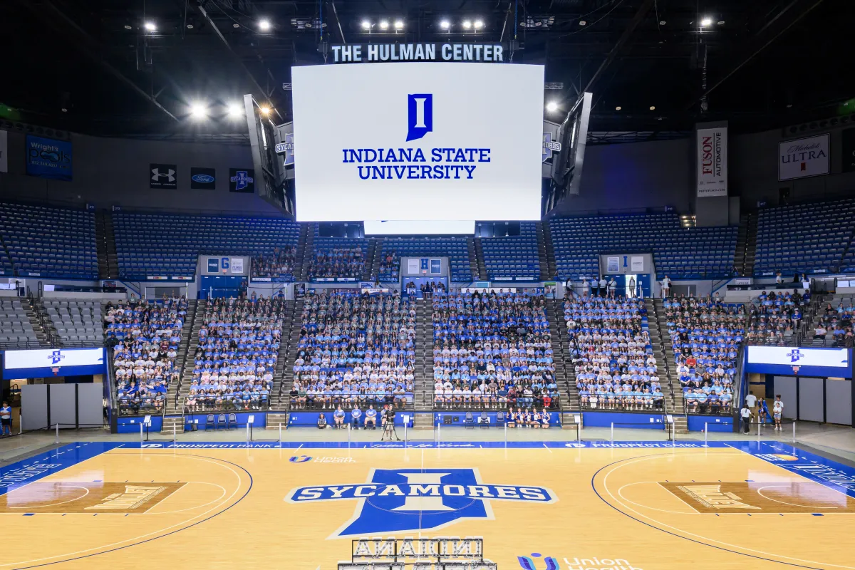 The image shows the interior of the Hulman Center at Indiana State University. It features a large basketball court with the Sycamores logo at center court. A big screen above displays the university's logo and name. The seating area is filled with people, most wearing white shirts.